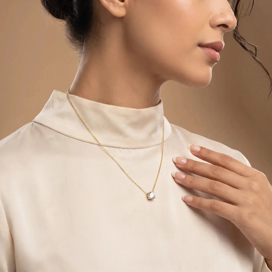 A close-up lifestyle shot of a model wearing a 1.05 CT Round Cut Diamond Chain Pendant, styled with a cream-colored mock neck top against a warm beige background.