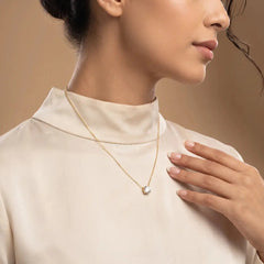 A close-up lifestyle shot of a model wearing a 1.05 CT Round Cut Diamond Chain Pendant, styled with a cream-colored mock neck top against a warm beige background.