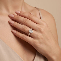 A close-up lifestyle shot of a woman's hand wearing a silver-toned halo ring with a clear center stone and small side stones, set against a soft beige background.
