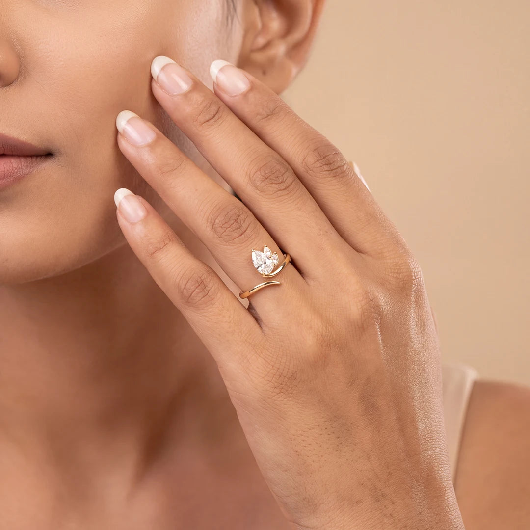 A close-up lifestyle shot of a woman's hand wearing a delicate yellow gold ring with a small white Pear-shaped diamond, set against a soft beige background.